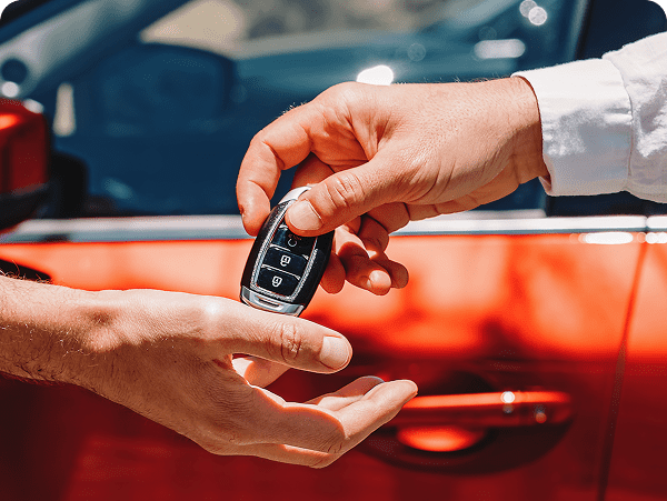 Person handing over a car key fob to another near a red car.